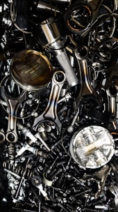 A chaotic assortment of various metal engine parts, including pistons, connecting rods, nuts, bolts, and springs. The components have a metallic sheen, and some show signs of wear and oxidation, suggesting they are used or discarded.