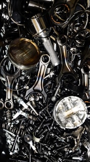 A chaotic assortment of various metal engine parts, including pistons, connecting rods, nuts, bolts, and springs. The components have a metallic sheen, and some show signs of wear and oxidation, suggesting they are used or discarded.
