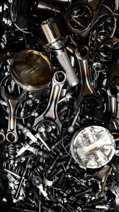 A chaotic assortment of various metal engine parts, including pistons, connecting rods, nuts, bolts, and springs. The components have a metallic sheen, and some show signs of wear and oxidation, suggesting they are used or discarded.