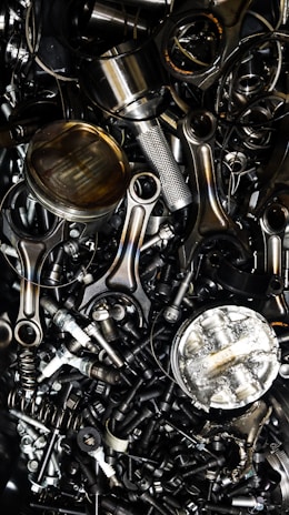 A chaotic assortment of various metal engine parts, including pistons, connecting rods, nuts, bolts, and springs. The components have a metallic sheen, and some show signs of wear and oxidation, suggesting they are used or discarded.