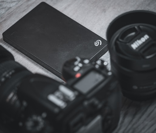 A black external hard drive is placed on a wooden surface surrounded by a camera and a camera lens. The lens cap has the Nikon logo, and the hard drive has a Seagate logo.