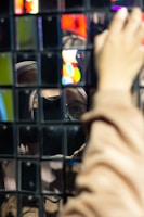 A reflection of a person wearing glasses and a hood is captured in a wall of mirrored tiles. The reflected image appears distorted and fragmented due to the multiple square mirrors. A hand is visible reaching toward the surface, adding depth to the image. Vibrant neon colors are reflected, suggesting a lively or urban setting.