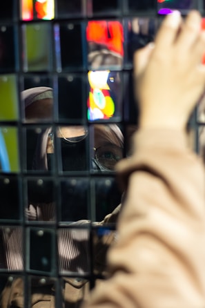 A reflection of a person wearing glasses and a hood is captured in a wall of mirrored tiles. The reflected image appears distorted and fragmented due to the multiple square mirrors. A hand is visible reaching toward the surface, adding depth to the image. Vibrant neon colors are reflected, suggesting a lively or urban setting.