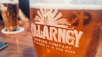 A close-up view of a glass filled with beer from Killarney Brewing Company, featuring a golden amber color. The frothy head is visible at the top of the glass, with the company's logo and slogan printed prominently in white on the glass surface. In the background, another similar glass of beer is slightly blurred, resting on a table.
