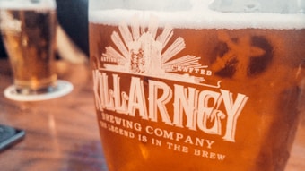 A close-up view of a glass filled with beer from Killarney Brewing Company, featuring a golden amber color. The frothy head is visible at the top of the glass, with the company's logo and slogan printed prominently in white on the glass surface. In the background, another similar glass of beer is slightly blurred, resting on a table.