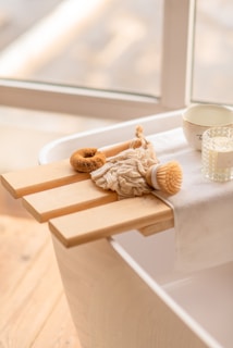 A serene spa-like setting with a wooden bath tray placed across a bathtub. The tray holds a loofah, a textured cloth, a natural bristle brush, a small bowl, and a glass candle. The soft lighting and neutral color scheme enhance the calming ambiance.