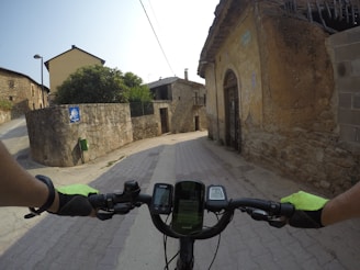 A cyclist's view as they ride along a narrow, paved street bordered by stone buildings. The cyclist's hands and handlebars are prominent in the foreground, with GPS and cycling data devices attached. The street features old architecture with rustic, browning walls and a few signs. A small greenery can be seen on the left, adding contrast to the stone structures.