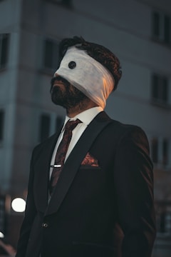 A man in a formal suit stands outdoors, wearing a blindfold that appears to be made of white fabric with a button detail. The background is an urban setting, likely a building at dusk or night. The man's facial hair and neatly styled hair contribute to his sophisticated appearance.