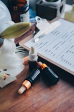 A wooden tray holds several essential oil bottles with droppers, accompanied by a white flower. In the background, there is a clipboard with a form partially visible, along with a small potted plant and an open brown bottle.