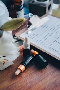 A wooden tray holds several essential oil bottles with droppers, accompanied by a white flower. In the background, there is a clipboard with a form partially visible, along with a small potted plant and an open brown bottle.
