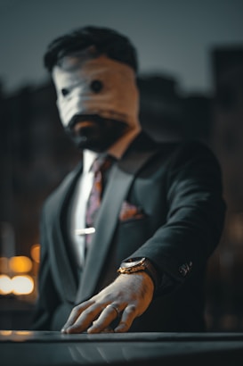 A person in a suit with a bandaged head, covering their face completely, is reaching forward with their hand. A watch and ring are visible on their wrist and finger. The background is blurred with bokeh lights.