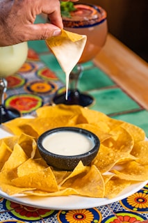 A hand is holding a tortilla chip dipped in a creamy white sauce, with a plate of chips surrounding a bowl of the same sauce on a colorful table. In the background, there are two drinks in glasses with decorative rims.