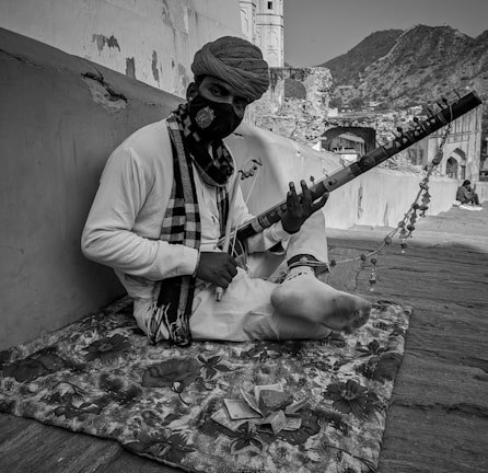 A person wearing traditional attire and a mask is sitting on a patterned mat with a stringed instrument. The background shows a scenic outdoor setting with architectural elements and distant hills.