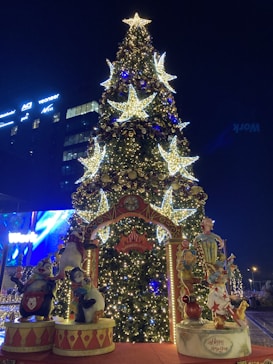 A large, decorated Christmas tree stands illuminated in the night with glowing star-shaped lights and ornaments. In front of the tree, there is an elaborate archway with colorful characters and decorations themed around a festive and whimsical setting, with 'wonderland' and 'Happy New Year' signs visible.