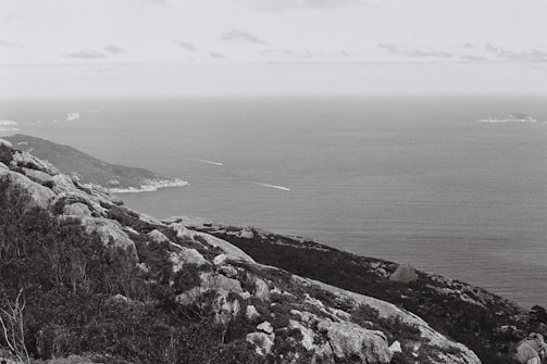 A black and white photograph captures a serene coastal landscape. In the foreground, rugged rocky terrain and vegetation create a textured scene, while the background reveals an expansive ocean view stretching towards the horizon. Small islands are visible in the distance, and the overall composition conveys a sense of vastness and tranquility.