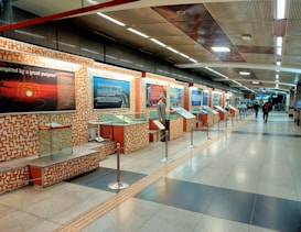 A modern metro station interior with an exhibition on display. The walls are adorned with informational boards and glass display cases. The ceiling features a series of lined panels with bright lighting. A few people walk through the spacious, well-lit corridor.
