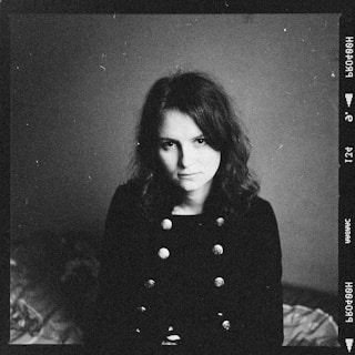 A black and white photograph featuring a person with shoulder-length hair, wearing a dark coat with distinctive buttons. The backdrop is plain and slightly textured. The image has visible grain and scratches, giving it a vintage or aged appearance.
