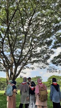 A group of people wearing scout scarves and backpacks gather under a large tree in a sunny park. The scene is vibrant with lush green grass and tree leaves. The individuals appear engaged in conversation, and the atmosphere is peaceful under the expansive blue sky with fluffy clouds.
