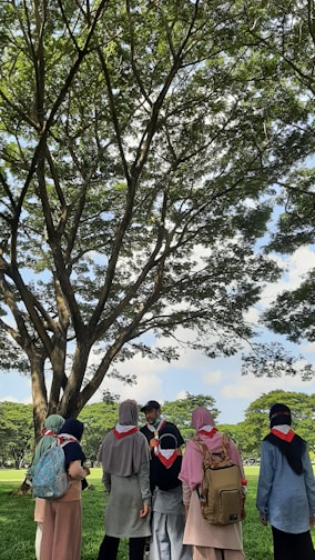 A group of people wearing scout scarves and backpacks gather under a large tree in a sunny park. The scene is vibrant with lush green grass and tree leaves. The individuals appear engaged in conversation, and the atmosphere is peaceful under the expansive blue sky with fluffy clouds.