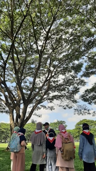 A group of people wearing scout scarves and backpacks gather under a large tree in a sunny park. The scene is vibrant with lush green grass and tree leaves. The individuals appear engaged in conversation, and the atmosphere is peaceful under the expansive blue sky with fluffy clouds.