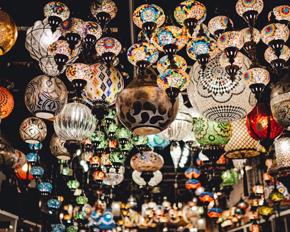 A vast array of colorful mosaic lamps hanging from the ceiling, each displaying intricate and diverse patterns. The setting suggests an artisanal marketplace or a specialty lighting store, with a warm and inviting atmosphere.