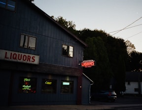A dimly lit street scene featuring a liquor store with blue walls and illuminated neon signs. Large red letters spell out 'LIQUORS' above the entrance, while additional neon signs are visible in the windows promoting brands like Tito's Handmade Vodka. The setting is near dusk with trees and a few residential buildings in the background.