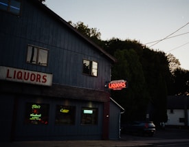 A dimly lit street scene featuring a liquor store with blue walls and illuminated neon signs. Large red letters spell out 'LIQUORS' above the entrance, while additional neon signs are visible in the windows promoting brands like Tito's Handmade Vodka. The setting is near dusk with trees and a few residential buildings in the background.