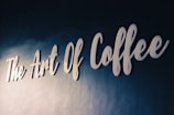 White cursive lettering spells out 'The Art Of Coffee' on a textured dark blue wall, conveying a sense of elegance and sophistication.