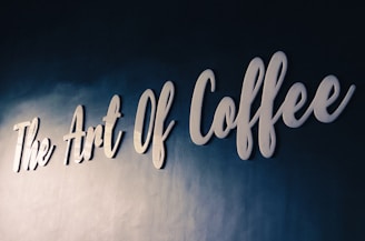 White cursive lettering spells out 'The Art Of Coffee' on a textured dark blue wall, conveying a sense of elegance and sophistication.