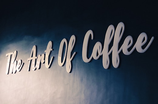 White cursive lettering spells out 'The Art Of Coffee' on a textured dark blue wall, conveying a sense of elegance and sophistication.