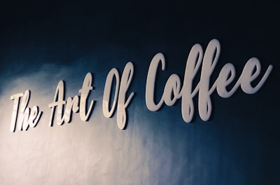 White cursive lettering spells out 'The Art Of Coffee' on a textured dark blue wall, conveying a sense of elegance and sophistication.