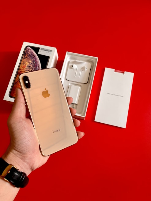 A hand holds a gold-colored iPhone with its back facing the camera. The iPhone is accompanied by its original packaging, which includes a box with a photo of the phone, a charger, and earphones laid out on a bright red surface.