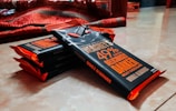 A stack of chocolate bars wrapped in dark packaging with bold orange and white text, resting on a smooth surface. The bars are aligned diagonally with padding visible on the edges. A red blanket is partially visible in the background.