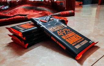 A stack of chocolate bars wrapped in dark packaging with bold orange and white text, resting on a smooth surface. The bars are aligned diagonally with padding visible on the edges. A red blanket is partially visible in the background.
