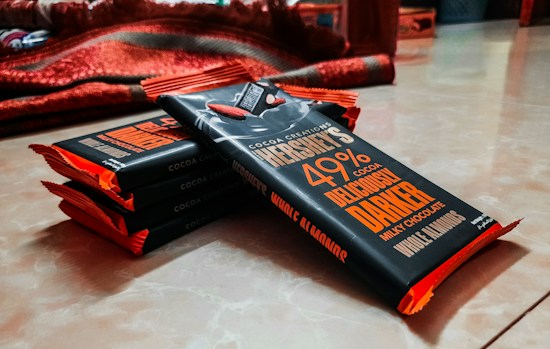 A stack of chocolate bars wrapped in dark packaging with bold orange and white text, resting on a smooth surface. The bars are aligned diagonally with padding visible on the edges. A red blanket is partially visible in the background.