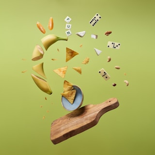 A dynamic levitation scene with objects seemingly floating in mid-air against a green background. Items include sliced bananas, orange segments, cheese pieces, tortilla chips, nuts, domino pieces, and Scrabble tiles spelling 'FUN'. The setup is centered over a wooden cutting board and a blue ceramic bowl.