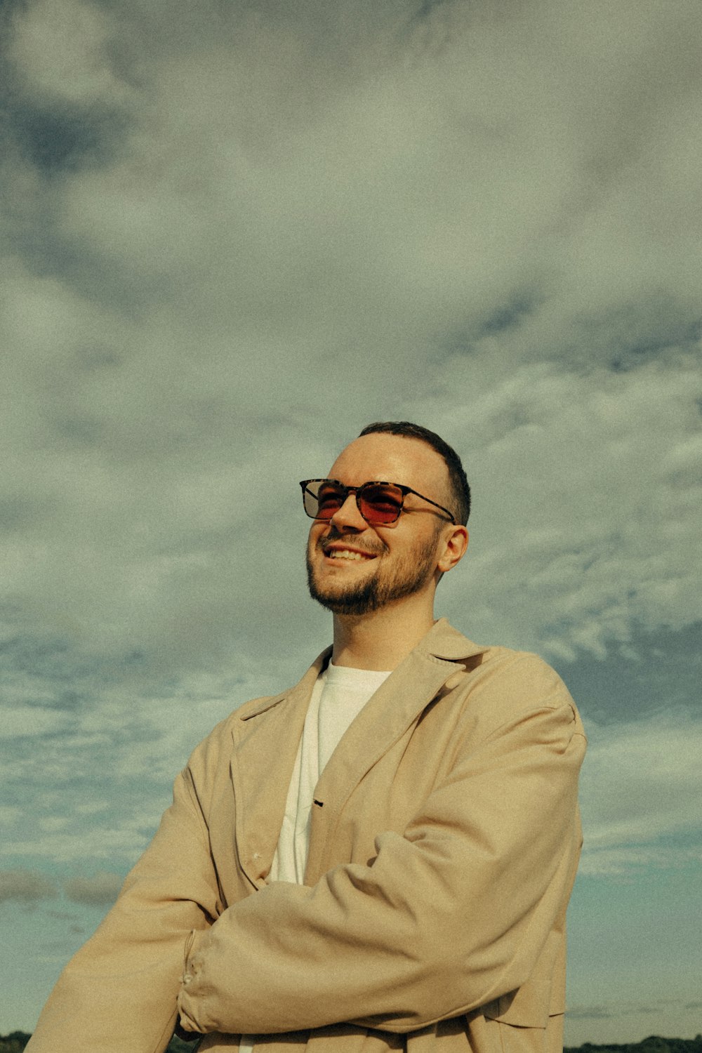 Un homme en trench-coat et lunettes de soleil debout devant un ciel nuageux