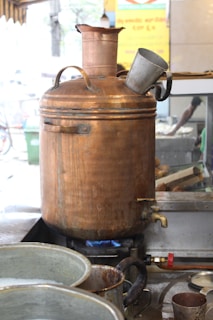 A large copper samovar is placed on a gas burner with a visible blue flame. The samovar is equipped with a spout for dispensing liquid, and a metal cup is resting on top. In the background, there are various kitchen utensils and a partially visible sign with text.