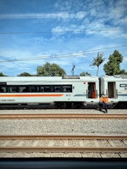 Two railway workers in orange safety vests stand near a stopped train on a railway track. The train has a white exterior with a horizontal orange stripe. Several railroad tracks are visible in the foreground, and the background includes trees and a partly cloudy sky.