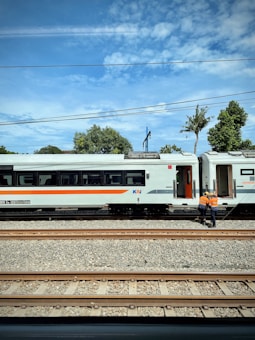Two railway workers in orange safety vests stand near a stopped train on a railway track. The train has a white exterior with a horizontal orange stripe. Several railroad tracks are visible in the foreground, and the background includes trees and a partly cloudy sky.