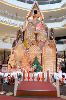 A large, elaborate gingerbread house display situated in a shopping mall, decorated with detailed patterns and colorful icing. Surrounding the gingerbread structure are decorated Christmas trees with baubles. A small red carpeted platform and a picket fence encircle the display, which is set against a background of modern architectural elements.