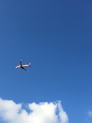 An orange passenger plane soaring above clouds with a clear blue sky background.