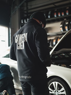 A person wearing a black jacket with 'CAMERA CREW' written on the back stands in a garage next to a white car with an open hood. Tires are neatly arranged on racks in the background, and another individual appears to be working near the vehicle.