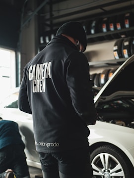 A person wearing a black jacket with 'CAMERA CREW' written on the back stands in a garage next to a white car with an open hood. Tires are neatly arranged on racks in the background, and another individual appears to be working near the vehicle.