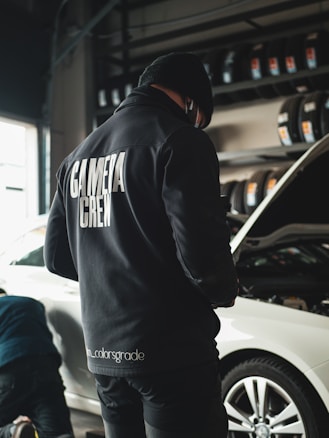 A person wearing a black jacket with 'CAMERA CREW' written on the back stands in a garage next to a white car with an open hood. Tires are neatly arranged on racks in the background, and another individual appears to be working near the vehicle.