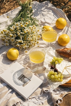 A picnic setting on a striped blanket includes a bouquet of white daisies, several lemons, two glasses of yellow juice, a loaf of bread, a wrapped cheese, and a book with a cover image of the Arc de Triomphe in Paris. There is also a bunch of grapes with a mix of green and dark purple grapes.