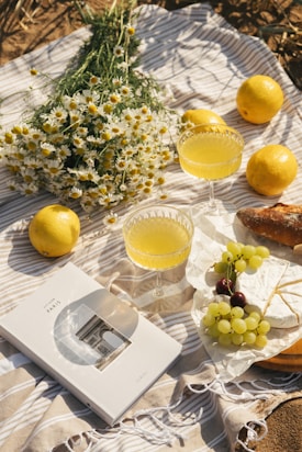 A picnic setting on a striped blanket includes a bouquet of white daisies, several lemons, two glasses of yellow juice, a loaf of bread, a wrapped cheese, and a book with a cover image of the Arc de Triomphe in Paris. There is also a bunch of grapes with a mix of green and dark purple grapes.
