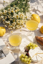 A picnic setup featuring two glasses of yellow lemonade alongside a bouquet of chamomile flowers on a striped blanket. Fresh lemons, a loaf of rustic bread, a wheel of brie cheese, green grapes, and cherries are also present. An open book lies nearby, contributing to a serene and leisurely atmosphere.