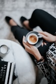 A person sits comfortably, holding a small cup of espresso in one hand. The individual wears dark pants and a stylish patterned blouse, accessorized with a watch. Nearby, a table holds stacked fashion books and a lit candle, adding a touch of elegance to the setting.