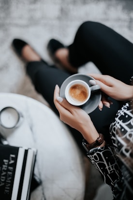 A person sits comfortably, holding a small cup of espresso in one hand. The individual wears dark pants and a stylish patterned blouse, accessorized with a watch. Nearby, a table holds stacked fashion books and a lit candle, adding a touch of elegance to the setting.